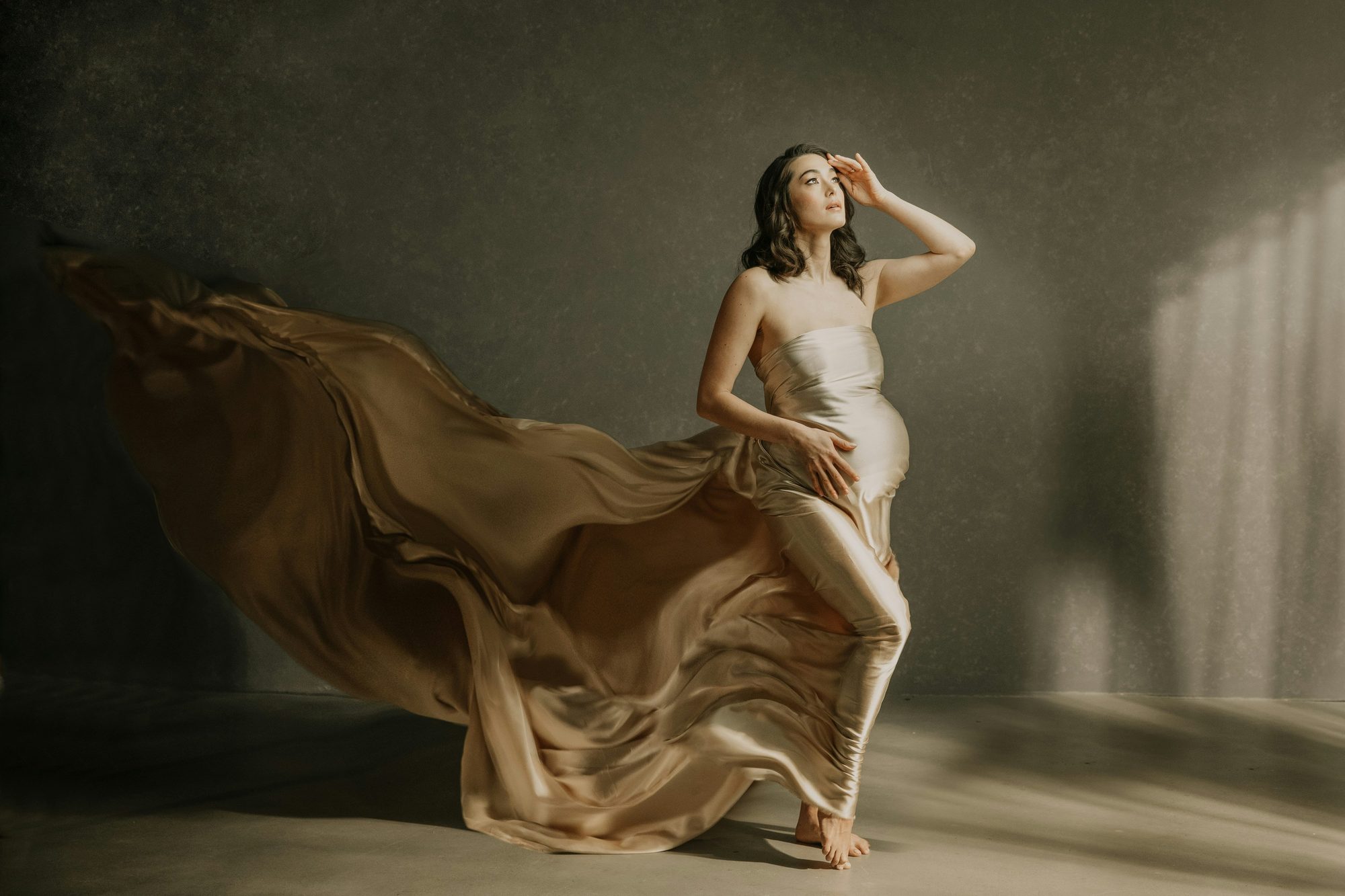 Pregnant woman wrapped in flowing champagne silk, standing barefoot in a shaft of window light against a dark wall.