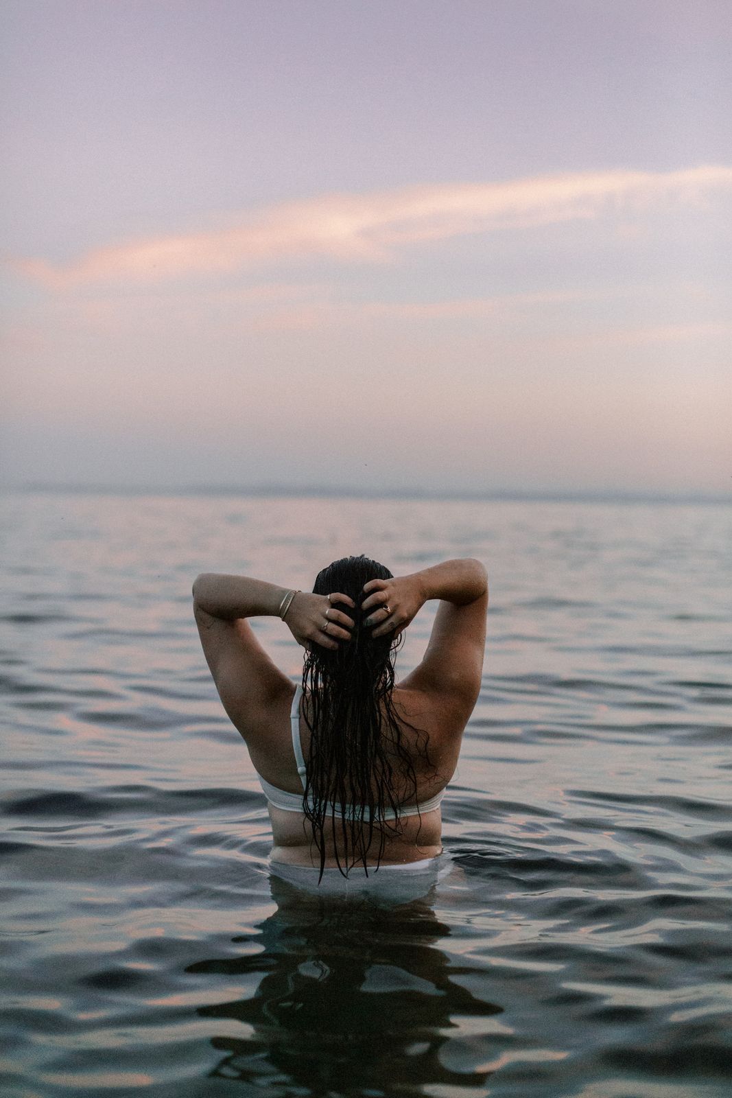 A woman standing in the sea at dusk, lifting her wet hair — a quiet portrait of a body at ease.